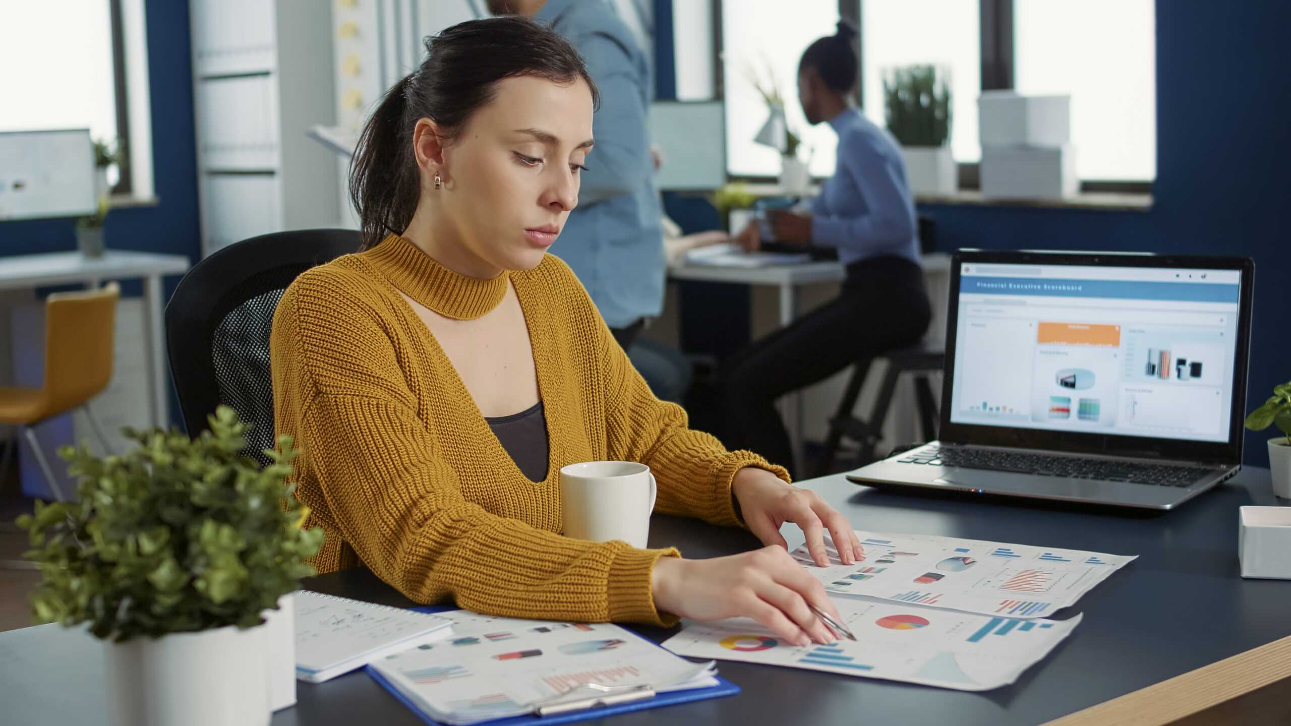 portrait startup employee comparing two sheets paper with charts analyzing financial data while drinking morning coffee woman working busy marketing department sitting desk with laptop 1 scaled